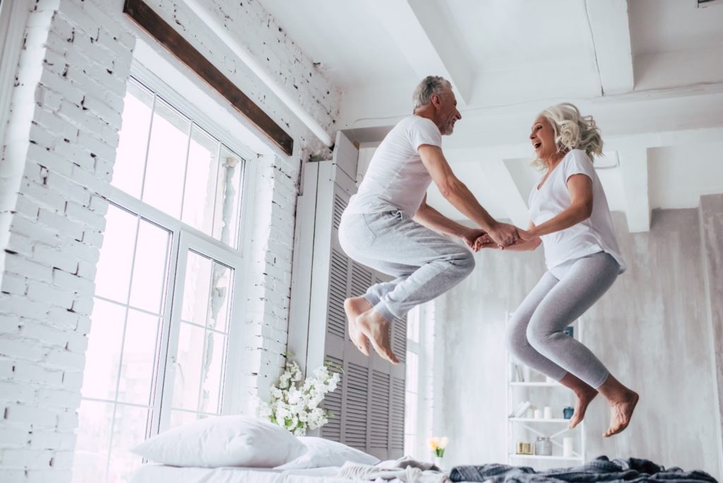 An older couple jumping happily on a bed holding hands.