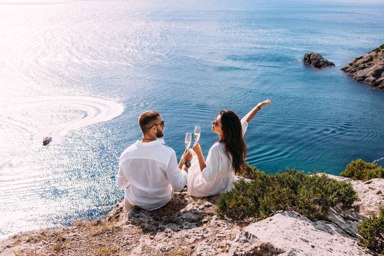 A man and woman happily sitting on a cliff overlooking the ocean drinking champagne for their honeymoon for the bella mansions wedding venue blog